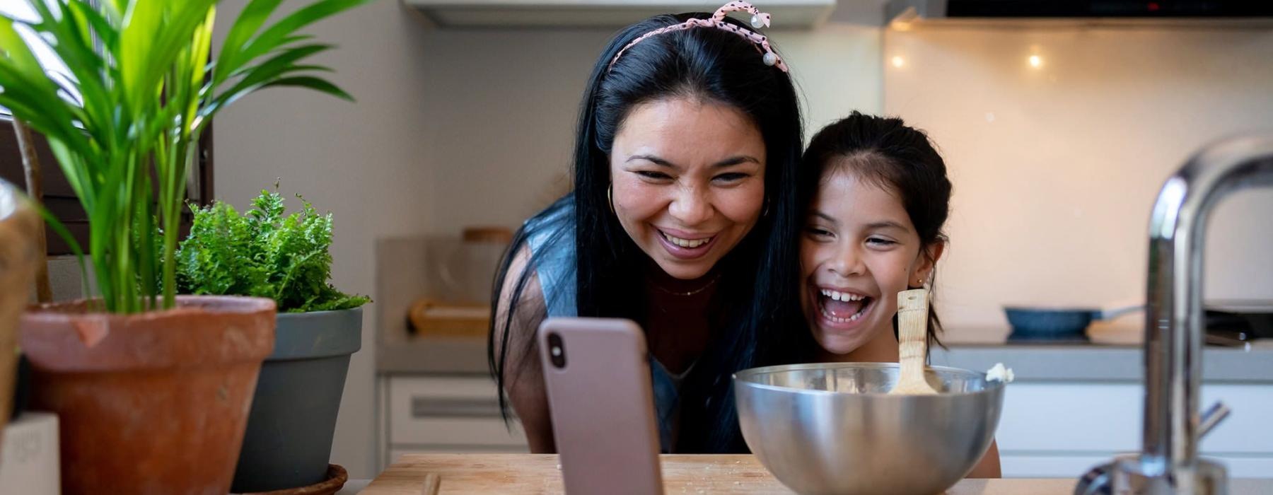 mother and young daughter smile at their phone as they cook in the kitchen